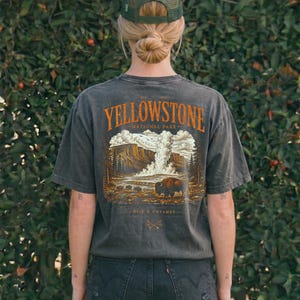 May include: Dark gray t-shirt featuring a Yellowstone National Park graphic with a bison and landscape illustration. The shirt has short sleeves and a crew neck. The text "Yellowstone National Park" is in orange. The person is wearing a green mesh cap and black denim shorts.