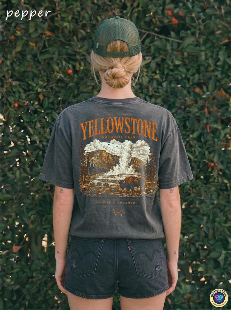 May include: Dark gray t-shirt featuring a Yellowstone National Park graphic with a bison and landscape illustration. The shirt has short sleeves and a crew neck. The text "Yellowstone National Park" is in orange. The person is wearing a green mesh cap and black denim shorts.