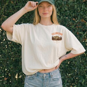 May include: Ivory-colored t-shirt with a Yellowstone National Park graphic. The shirt is paired with denim shorts and a green cap featuring a bear and mountain patch. The overall style is casual and outdoorsy.