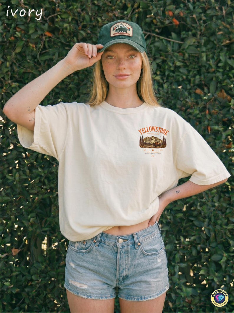 May include: Ivory-colored t-shirt with a Yellowstone National Park graphic. The shirt is paired with denim shorts and a green cap featuring a bear and mountain patch. The overall style is casual and outdoorsy.