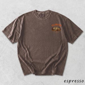 May include: Espresso-colored t-shirt with a crew neck and short sleeves. The shirt features a graphic on the upper chest with the word "Yellowstone" above a mountain and tree scene. The shirt has a vintage, worn look.