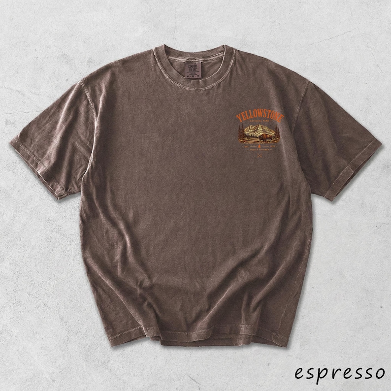 May include: Espresso-colored t-shirt with a crew neck and short sleeves. The shirt features a graphic on the upper chest with the word "Yellowstone" above a mountain and tree scene. The shirt has a vintage, worn look.