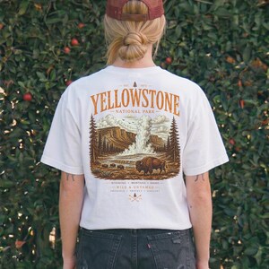 May include: White t-shirt featuring a Yellowstone National Park graphic. The design includes a bison, geyser, and text. The shirt is worn with black denim shorts and a maroon trucker hat. The text on the shirt reads "Yellowstone National Park".