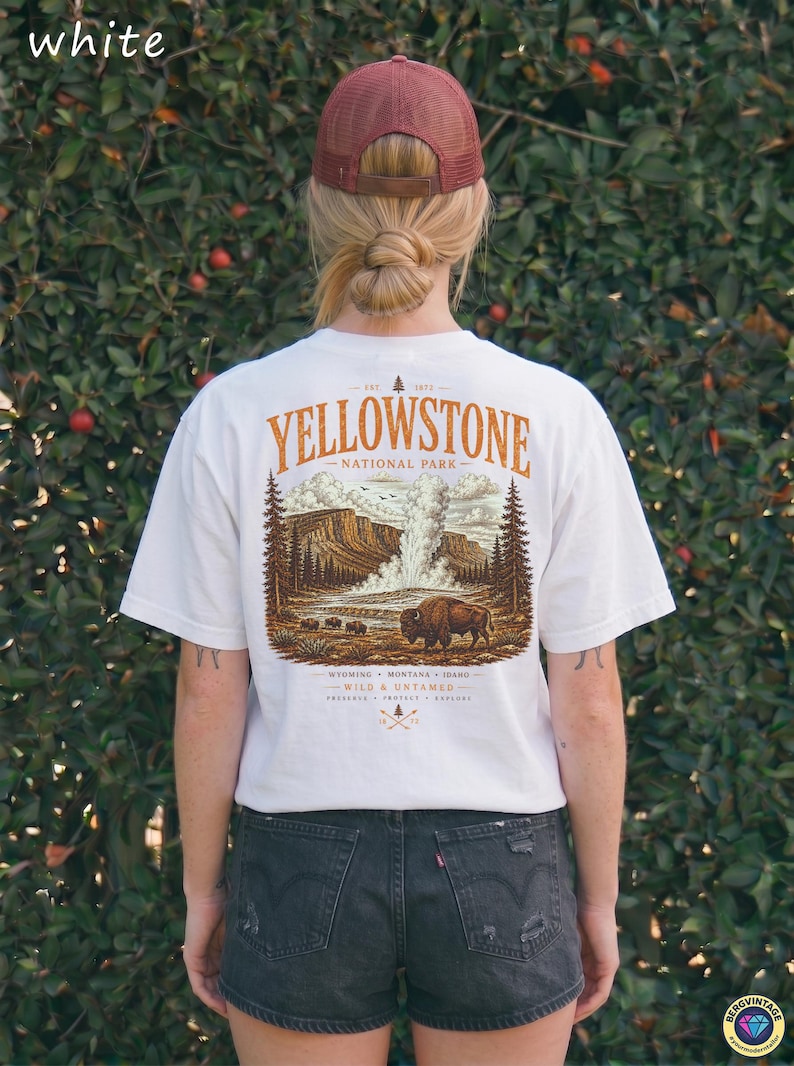 May include: White t-shirt featuring a Yellowstone National Park graphic. The design includes a bison, geyser, and text. The shirt is worn with black denim shorts and a maroon trucker hat. The text on the shirt reads "Yellowstone National Park".