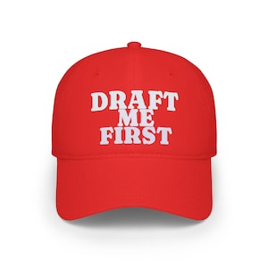 May include: A bright red baseball cap with the text "DRAFT ME FIRST" in bold white letters. The cap has a curved brim and a classic six-panel design, suitable for casual wear.