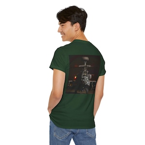 Puede incluir: Camiseta verde oscuro con una imagen detallada de un helicóptero en la espalda. La imagen del helicóptero está sobre un fondo oscuro, con luces visibles. La camiseta está hecha de un material suave.