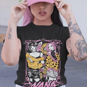 May include: Black t-shirt featuring a pink and yellow graphic of two anime-style women with the word "NANA" in a decorative font. The person in the image is wearing a pink baseball cap and blue jeans.