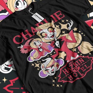 May include: Black t-shirt featuring a colorful graphic of two cartoon characters with the text "CHARLIE" and "HAZBIN HOTEL". The design includes stars and a decorative border. A white tag is attached to the shirt.
