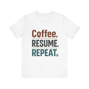 May include: White t-shirt with the text "Coffee. RESUME. REPEAT." in brown and teal lettering. The word "Coffee" has a small coffee cup icon above it. The shirt is made of a soft, comfortable material.