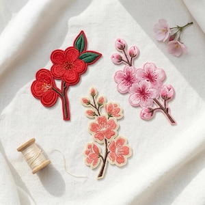 May include: Three embroidered flower patches featuring a red blossom with green leaves, a light pink cherry blossom branch with buds, and a coral-colored floral sprig. A small wooden spool of light-colored thread sits nearby on a white fabric surface.
