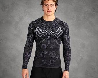 May include: A long-sleeve compression shirt featuring a black and grey spiderweb pattern across the chest, back, and sleeves. The garment displays a prominent white, stylised spider emblem centred on the chest, set against a dark, textured fabric.