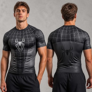 Op de afbeelding: Een zwart compressieshirt met korte mouwen, voorzien van een wit spinnenembleem op de borst en een zwart spinnenembleem op de rug. De bovenkant van de borst en de schouders hebben een wit spinnenwebpatroon op een zwarte ondergrond.