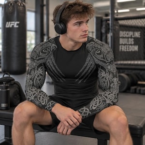 May include: A black long-sleeve compression shirt featuring intricate grey Celtic-inspired knotwork patterns on the sleeves and shoulders, paired with black athletic shorts. The shirt has a crew neckline and a fitted design.
