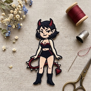 May include: Embroidered patch featuring a cartoon devil woman with black hair, red horns, and a matching tail. She wears a black outfit with red accents. The patch is surrounded by sewing supplies and dried flowers.