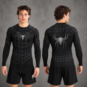 May include: A black long-sleeve compression shirt featuring a white spider web pattern across the chest and shoulders, with a large silver spider graphic centred on the front and back, paired with plain black athletic shorts.
