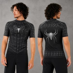 May include: A black short-sleeve compression shirt featuring a white spider web pattern across the chest and shoulders, with a large silver spider emblem centered on the front and back. The design includes a grid-like web detail on the hem.