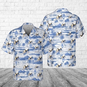 May include: A short-sleeved, button-down shirt with a tropical print featuring a repeating pattern of cartoon characters, palm trees, sailboats, and ocean waves. The shirt is primarily light blue and white, with a V-neck collar and a pocket on the chest.