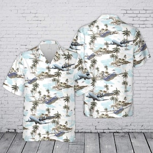 Puede incluir: Una camisa de manga corta con botones que presenta un patr&oacute;n repetitivo de aviones grises volando sobre un paisaje de palmeras y agua azul claro. La camisa tiene un cuello cl&aacute;sico, una tapeta de botones frontal y un color base blanco.
