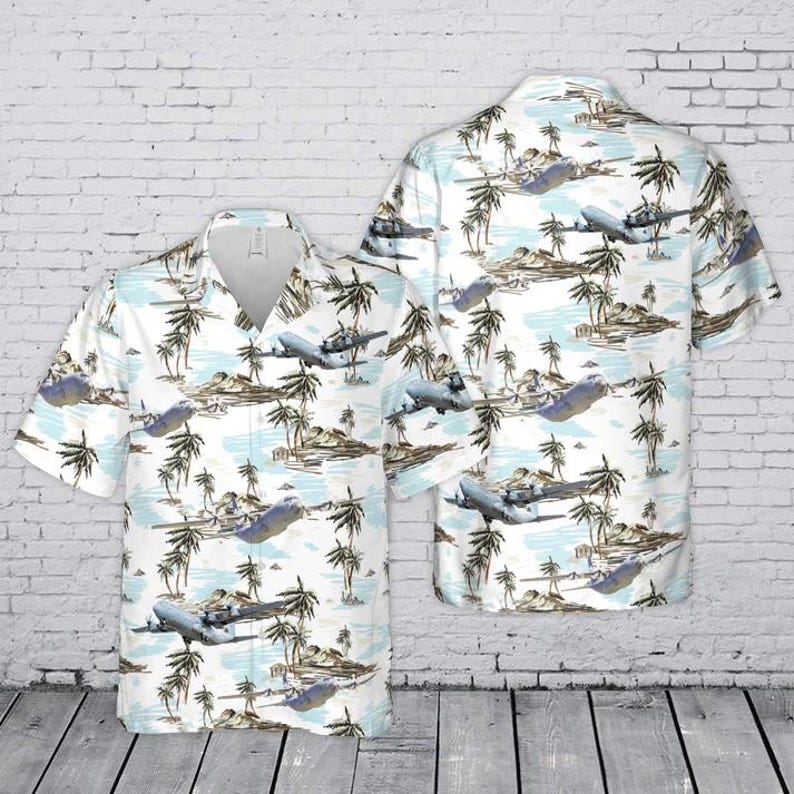 Puede incluir: Una camisa de manga corta con botones que presenta un patr&oacute;n repetitivo de aviones grises volando sobre un paisaje de palmeras y agua azul claro. La camisa tiene un cuello cl&aacute;sico, una tapeta de botones frontal y un color base blanco.