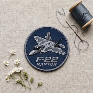May include: A navy blue embroidered patch featuring a detailed illustration of an F-22 Raptor fighter jet. The text "F-22 RAPTOR" is embroidered below the jet. The patch is round with a gray border. A spool of gray thread and a needle are also in the image.