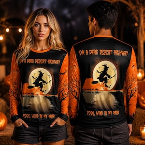 Könnte beinhalten: Ein Langarmshirt mit orangefarbenen Ärmeln und schwarzem Rumpf, das eine Hexe auf einem Besen vor einem Vollmond zeigt. Der Text ON A DARK DESERT HIGHWAY und COOL WIND IN MY HAIR ist in orangefarbenen Buchstaben auf der Vorder- und Rückseite gedruckt.