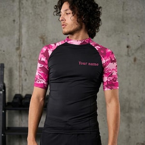 May include: Black and pink athletic top with short sleeves. The sleeves and collar have a pink camouflage design. The text "Your name" is printed in pink on the chest. Suitable for sports and exercise.