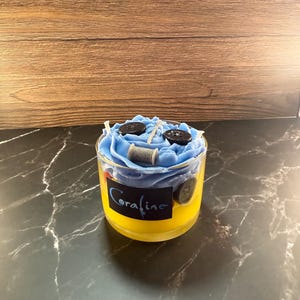 Peut inclure: Une bougie décorative dans un récipient en verre transparent. La bougie est jaune à la base, passant au bleu en haut, et comporte des boutons et une bobine de fil. Le mot "Coraline" est écrit sur le verre en script bleu.