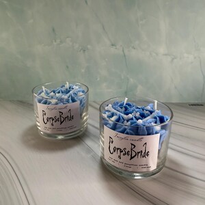 Peut inclure: Deux bougies en pot de verre remplies de fleurs de cire bleues et blanches. Chaque bougie porte une étiquette indiquant "Corpse Bride". Les bougies sont sur une surface en marbre blanc et gris.