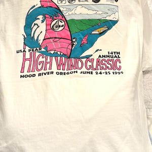 Puede incluir: Camiseta blanca con un gráfico colorido de un windsurfista sobre una ola, con una vela rosa y agua azul. El texto "HIGH WIND CLASSIC" se muestra, junto con "HOOD RIVER OREGON JUNE 24-25 1995".