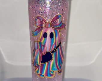 May include: A clear tumbler filled with pink glitter and a cartoon ghost design. The ghost is wearing a bow, carrying a drink, and a purse. The rim of the tumbler is decorated with blue rhinestones. A fun and whimsical drinkware item.