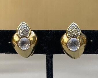 May include: A pair of gold-toned clip-on earrings. Each earring features a heart-shaped design with a large, clear, round crystal at the center and a smaller, teardrop-shaped cluster of crystals at the top. The earrings are displayed on a black velvet surface.
