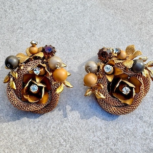 Puede incluir: Un par de pendientes de clip de tono dorado con un racimo de perlas falsas, diamantes de imitación y flores de metal.