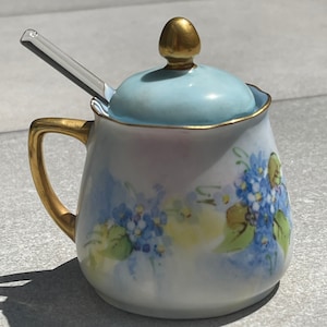 May include: A white porcelain sugar bowl with a blue and gold floral design. The bowl has a lid with a gold knob and a matching glass spoon.