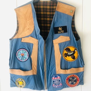 May include: A blue denim vest with brown leather accents and multiple patches. The patches include the words "Sportsman's Club", "Nodles Italy", "A.C. Schaf Maples, Italy", "NSSA", "Shooters Club of America Sponsored by Guns Magazine", and "United States Revolver Association".