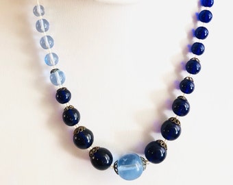 May include: A blue and clear glass bead necklace with a gradient of colour. The necklace is made of round beads with a silver accent on each bead. The necklace is on a white mannequin.