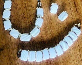Puede incluir: Un conjunto de collar, pulsera y pendientes de cerámica blanca con detalles dorados. La joyería presenta un diseño acanalado.