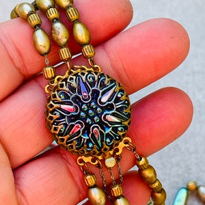 May include: A multi-strand necklace with gold-toned beads and a large, ornate pendant. The pendant features a circular, iridescent glass cabochon with a floral design.