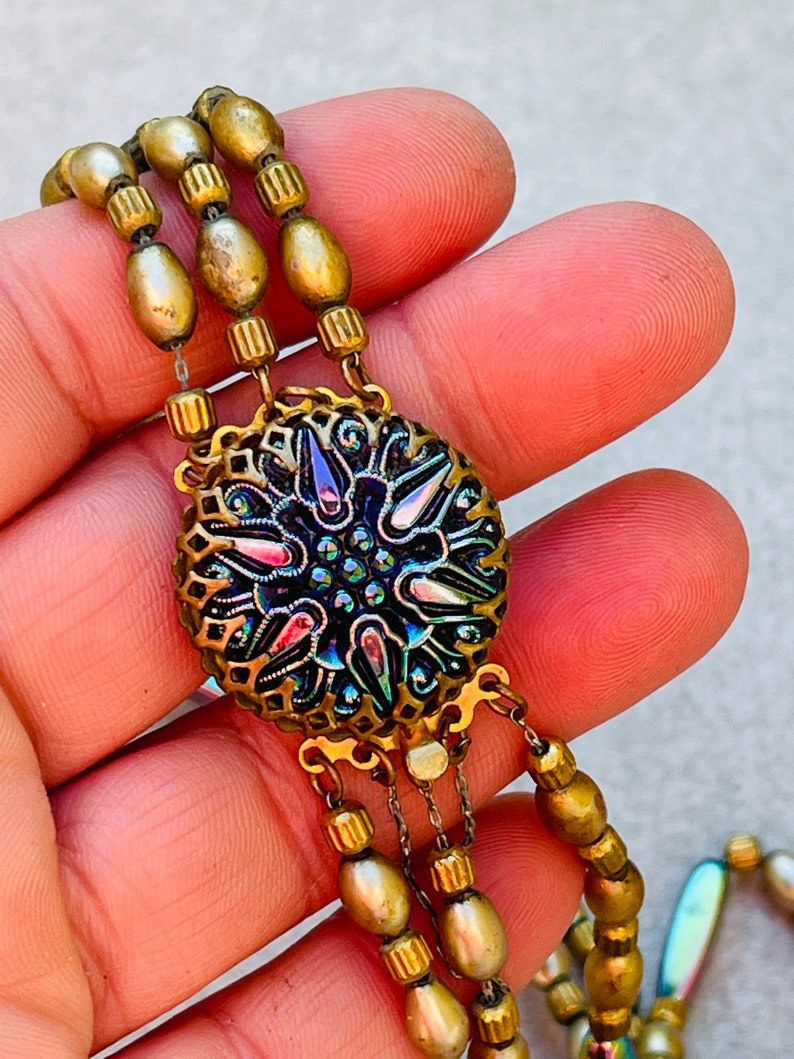 May include: A multi-strand necklace with gold-toned beads and a large, ornate pendant. The pendant features a circular, iridescent glass cabochon with a floral design.
