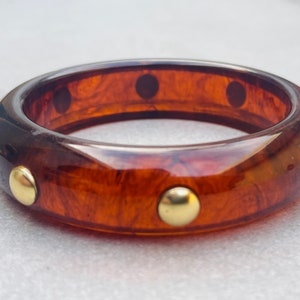 May include: A brown tortoiseshell bangle bracelet with gold-toned accents. The bracelet is made of plastic and has a smooth, glossy finish.