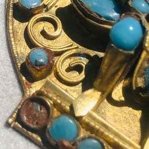 May include: A gold-toned metal brooch with a decorative design featuring blue turquoise stones. The brooch has a detailed floral pattern and intricate scrollwork.