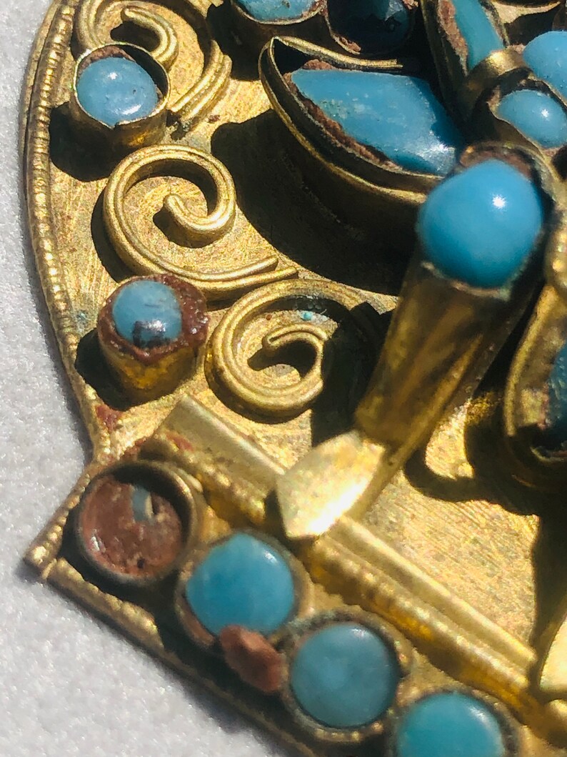 May include: A gold-toned metal brooch with a decorative design featuring blue turquoise stones. The brooch has a detailed floral pattern and intricate scrollwork.