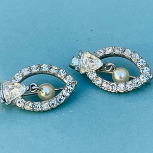 Könnte beinhalten: Ein Paar silberfarbene Ohrringe mit einer baumelnden Perle und einem Kristalldesign. Die Ohrringe sind oval geformt und haben eine Reihe von klaren Strasssteinen am Rand.