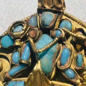 May include: A gold pendant with a turquoise stone and intricate details. The pendant is decorated with a crown and other decorative elements.