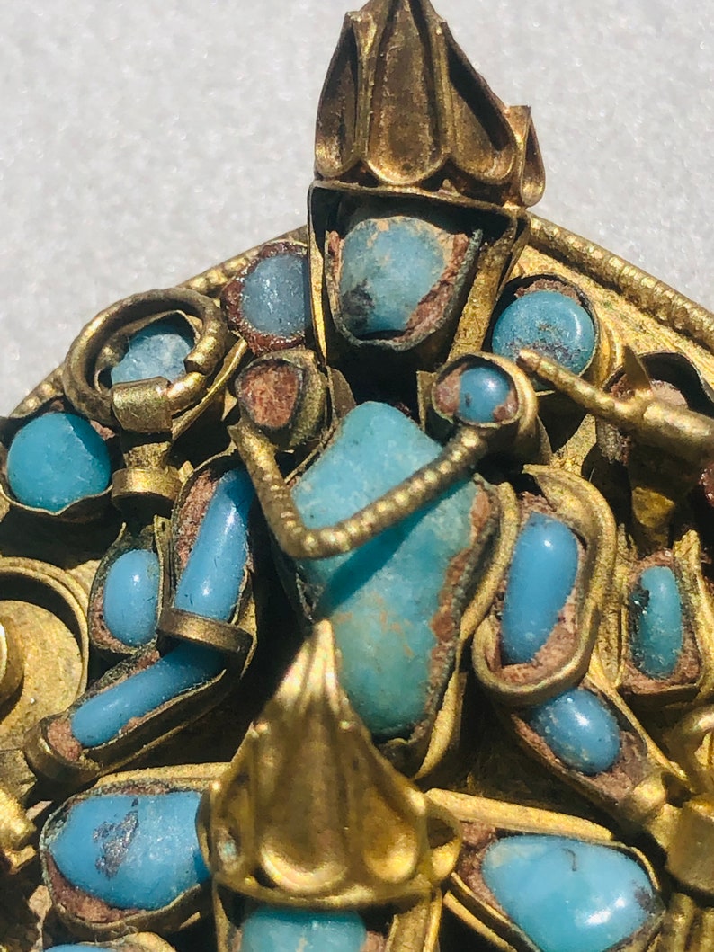 May include: A gold pendant with a turquoise stone and intricate details. The pendant is decorated with a crown and other decorative elements.