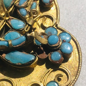 May include: A gold-toned pendant with a detailed design featuring blue turquoise stones. The pendant is shaped like a heart and has a decorative border.