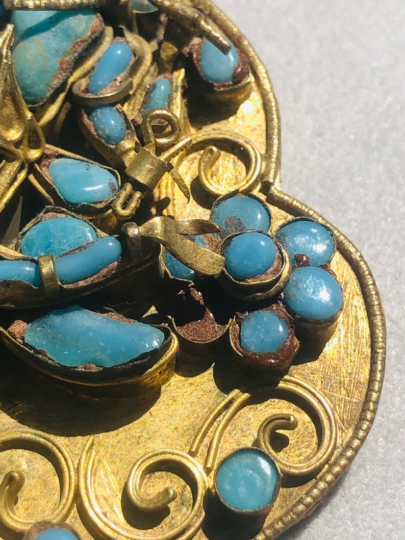 May include: A gold-toned pendant with a detailed design featuring blue turquoise stones. The pendant is shaped like a heart and has a decorative border.