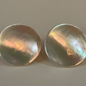 Pode incluir: Um par de brincos redondos e iridescentes. Os brincos têm um brilho perolado com toques de rosa, verde e dourado. Os brincos são um design simples e elegante.