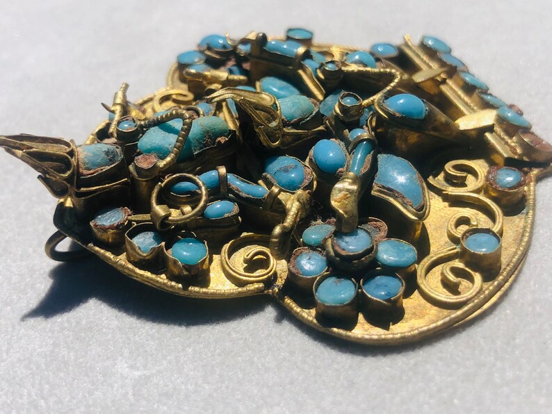 May include: A gold-toned pendant with a heart-shaped design, featuring intricate scrollwork and numerous turquoise stones.