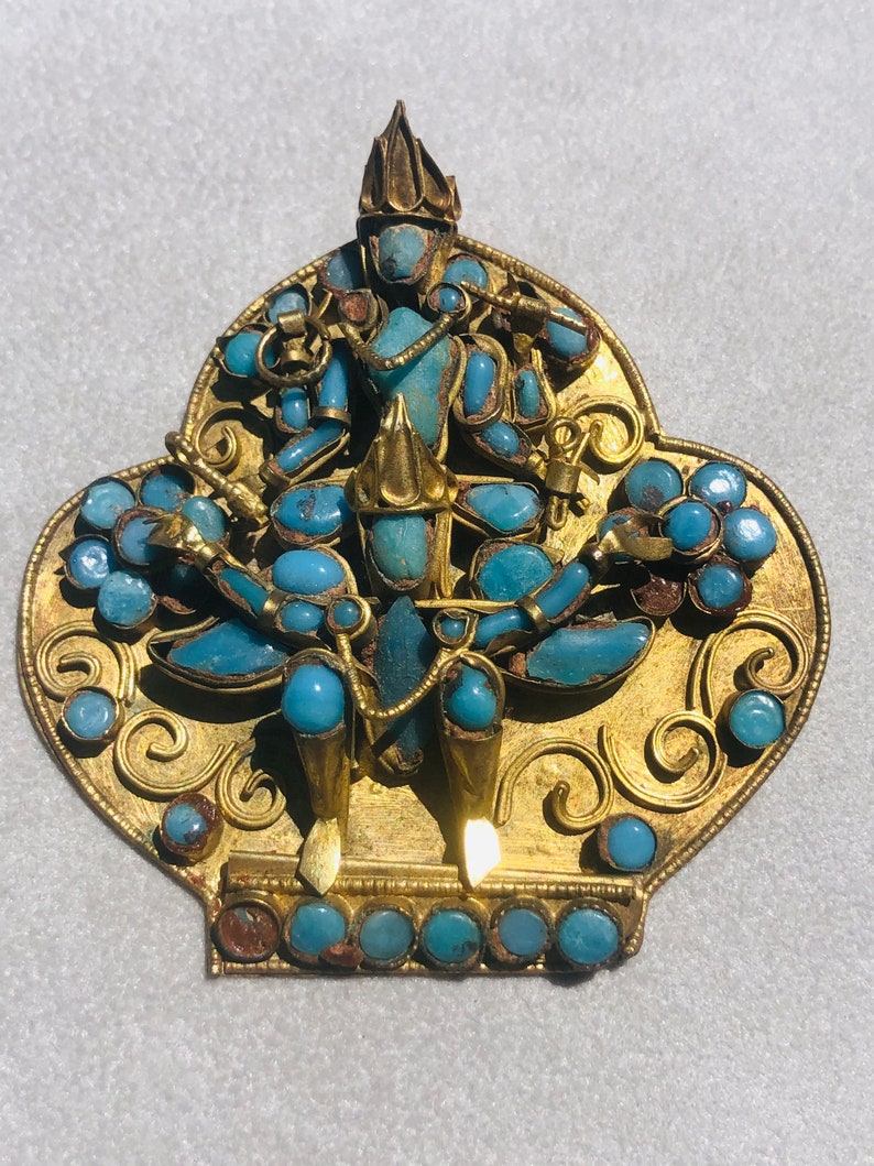May include: A gold pendant with a turquoise blue deity figure in the center. The pendant is decorated with intricate gold filigree and turquoise blue stones.