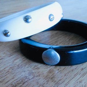 May include: Two bangle bracelets, one white and one black, with silver accents. The white bracelet has four silver screws on the top and the black bracelet has one large silver screw on the bottom.